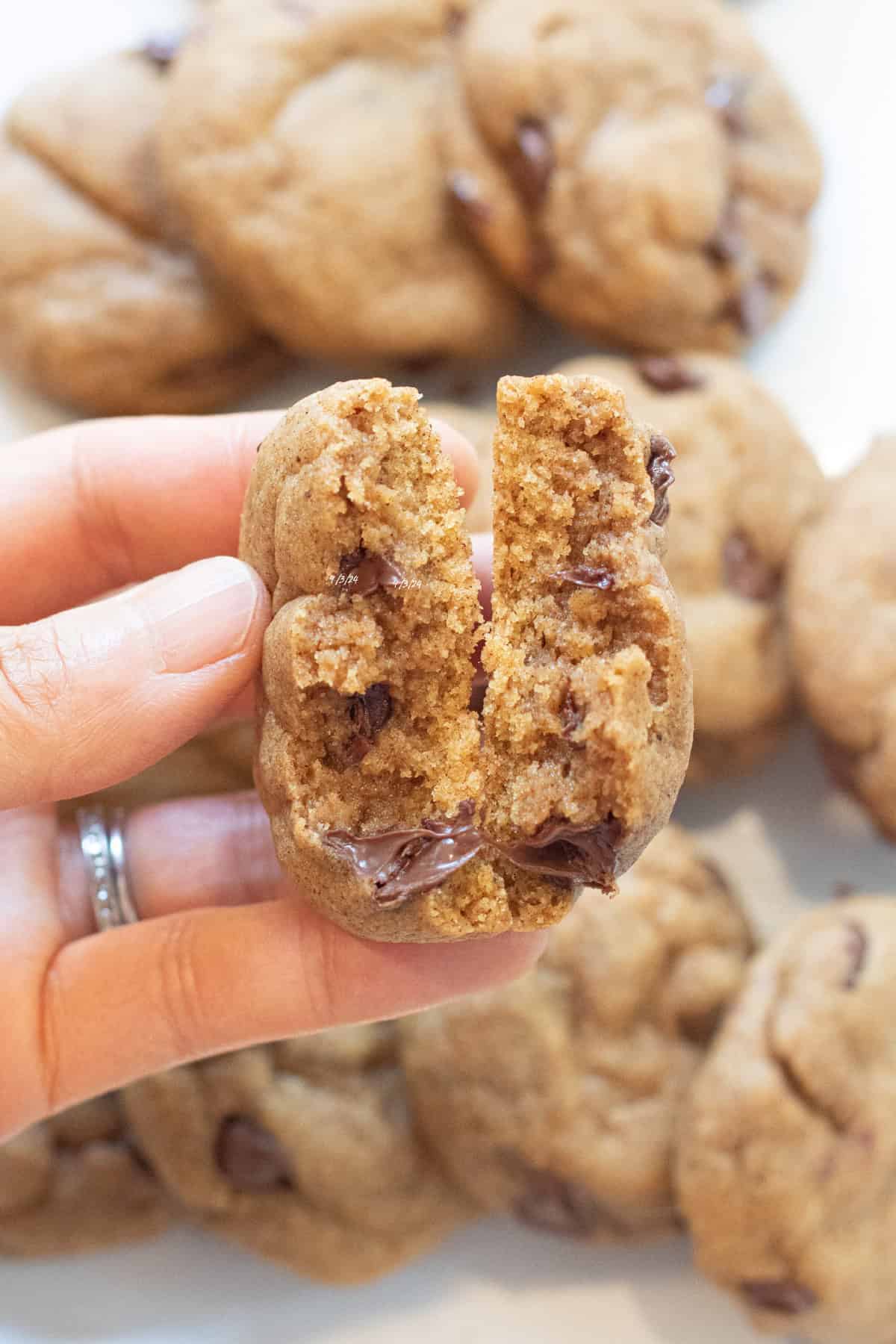 a hand holding a halved chocolate chip cookie over a plate of cookies.