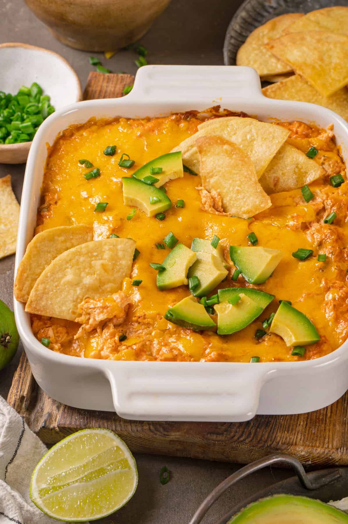Buffalo chicken dip (topped with tortilla chips, sliced avocado, lime slices, and chopped green onions) in a white baking dish.