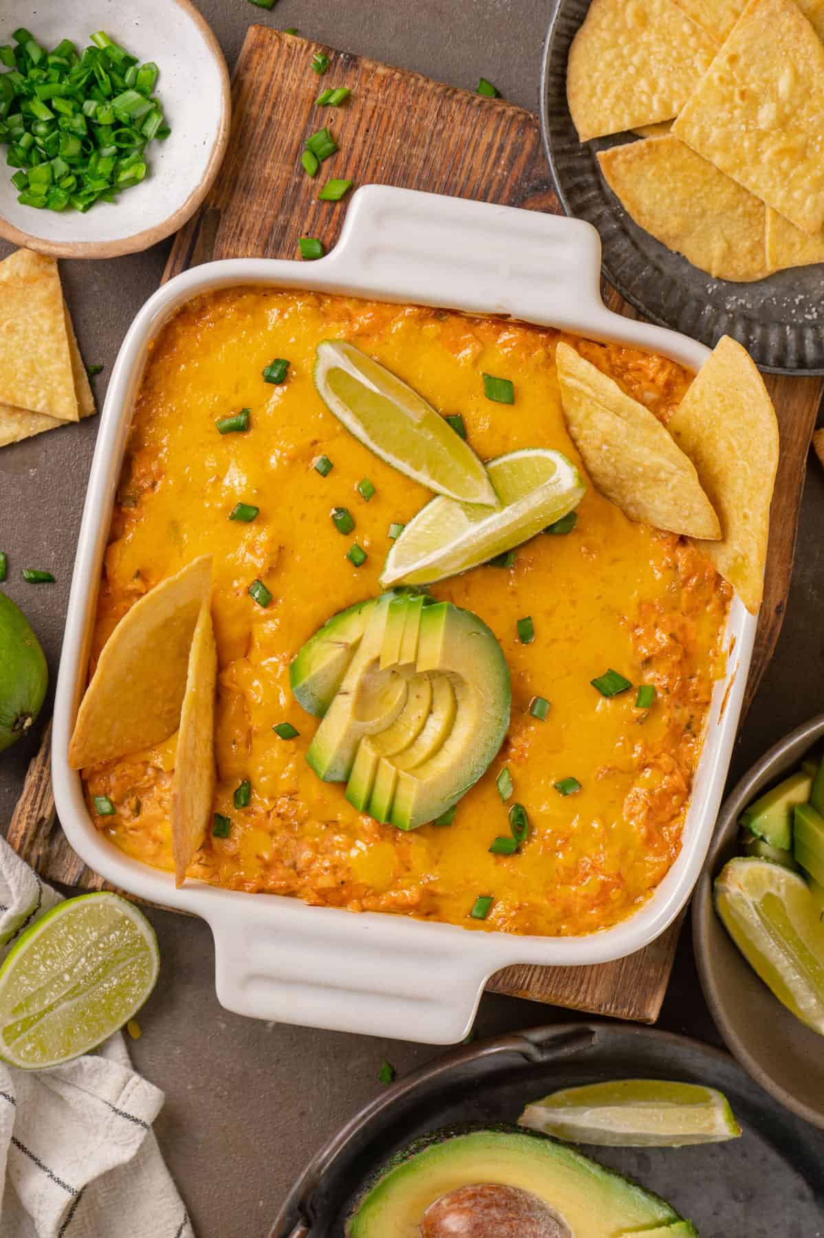 Buffalo chicken dip (topped with tortilla chips, sliced avocado, lime slices, and chopped green onions) in a white baking dish.