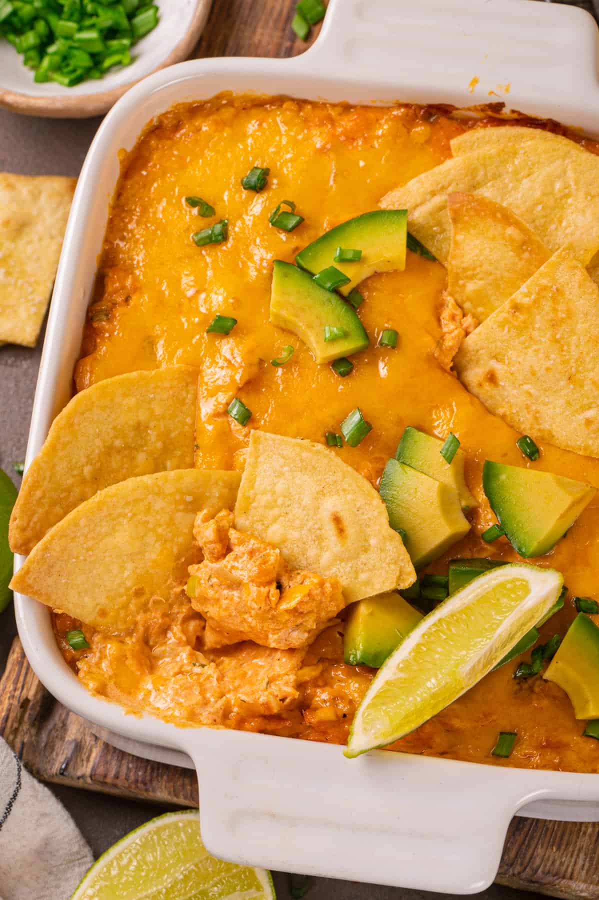 Buffalo chicken dip (topped with tortilla chips, sliced avocado, lime slices, and chopped green onions) in a white baking dish.