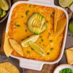 Buffalo chicken dip (topped with tortilla chips, sliced avocado, lime slices, and chopped green onions) in a white baking dish.
