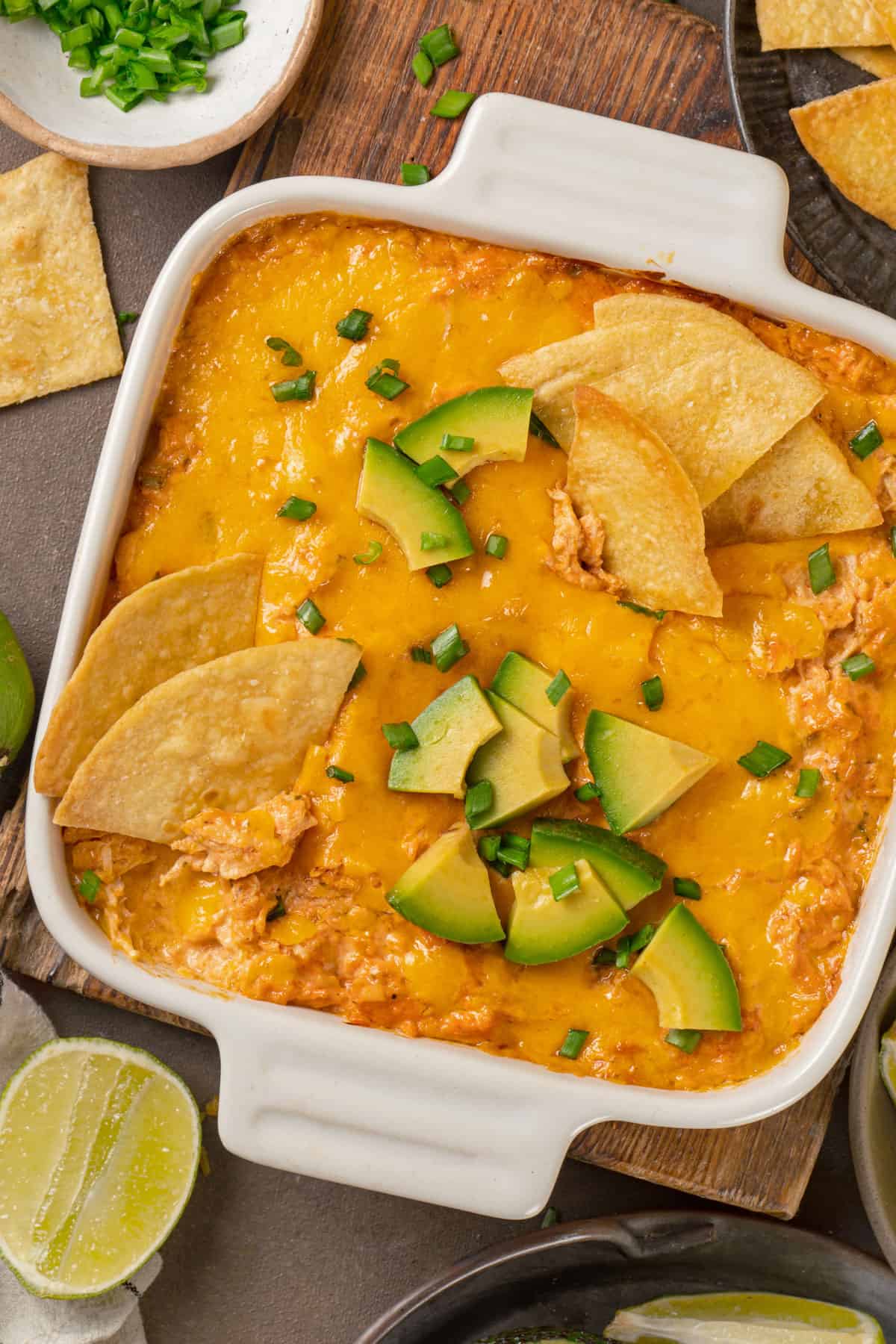 Buffalo chicken dip (topped with tortilla chips, sliced avocado, lime slices, and chopped green onions) in a white baking dish.