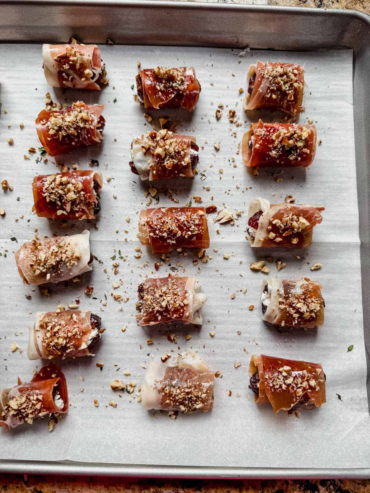 Prosciutto wrapped dates topped with pecans on a baking pan.